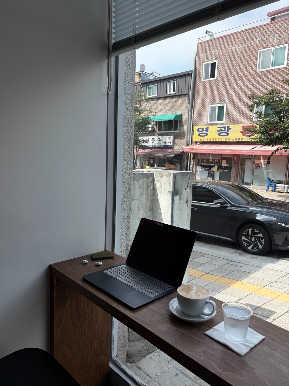 A workspace with a laptop and a warm Cafe Latte set on a bar table by a cafe window looking out onto Seochon 창밖으로 서촌 풍경이 보이는 카페 창가 바 테이블에 노트북과 따뜻한 카페라떼를 올려둔 작업 공간