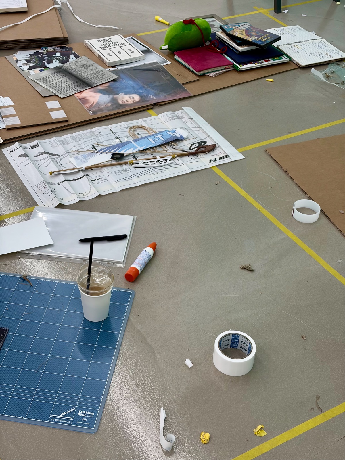 A workspace strewn with rubber-band-powered airplane blueprints, a cutting mat, and SeMA exhibition archive books 고무동력기 설계도와 커팅 매트, SeMA 전시 아카이브 책 등이 널려 있는 작업 공간