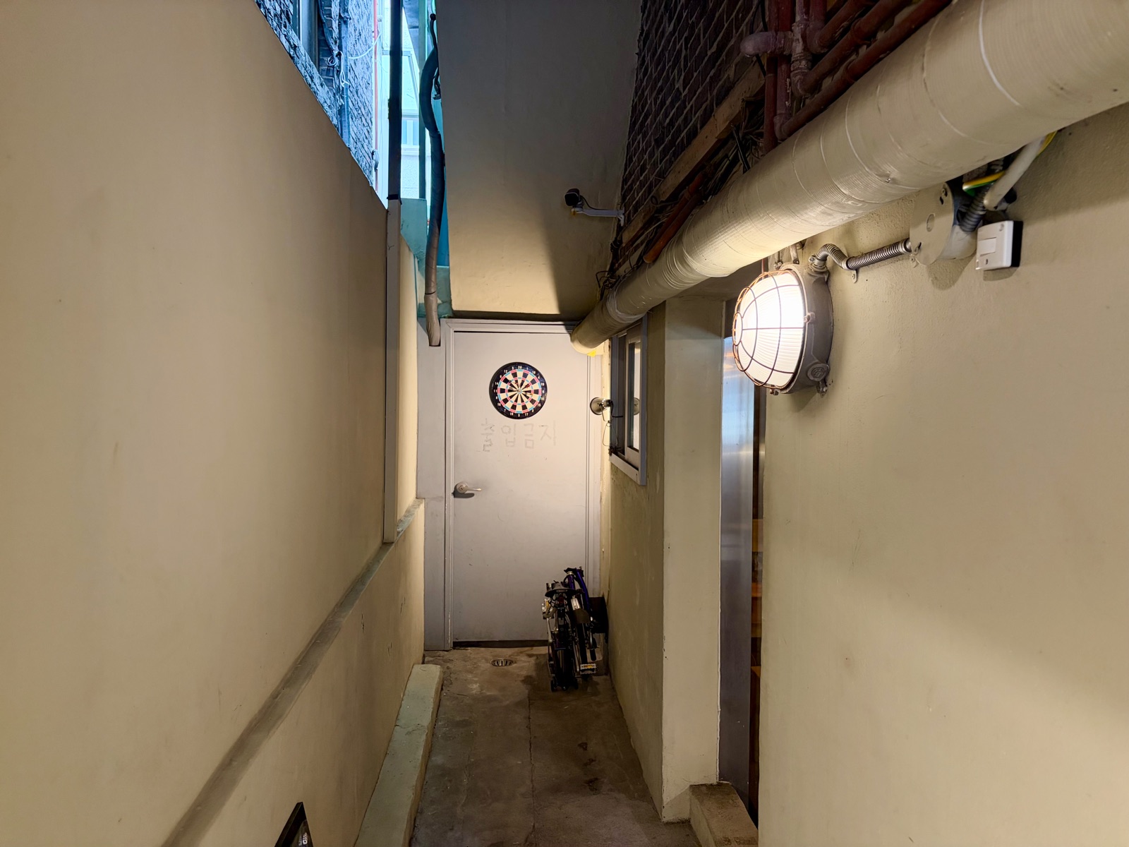 A narrow passageway entrance with a dartboard hanging on the door and a folding bicycle parked beside it 다트판이 걸린 문과 접이식 자전거가 세워진 좁은 통로-입구의 풍경