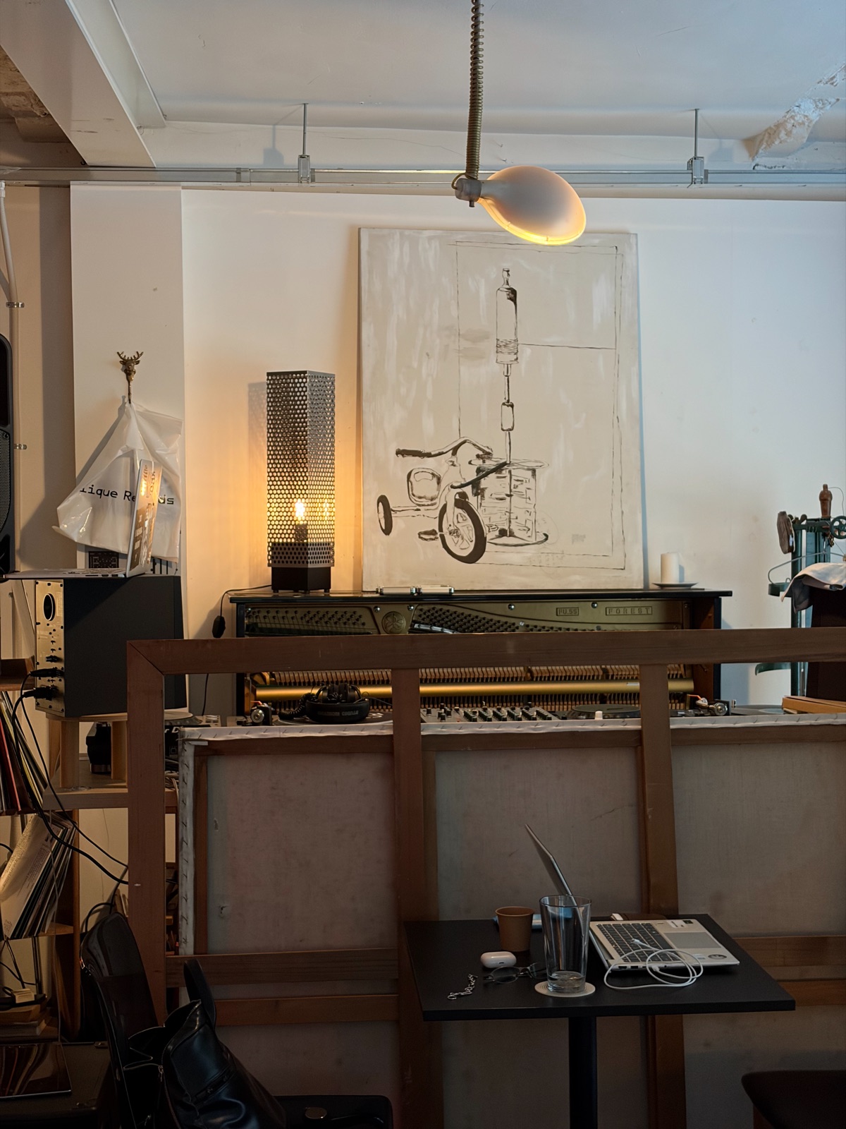 A cozy workspace with a piano and audio equipment visible beyond a small desk holding a laptop 노트북이 놓인 작은 책상 너머로 피아노와 음향 장비가 보이는 아늑한 작업 공간