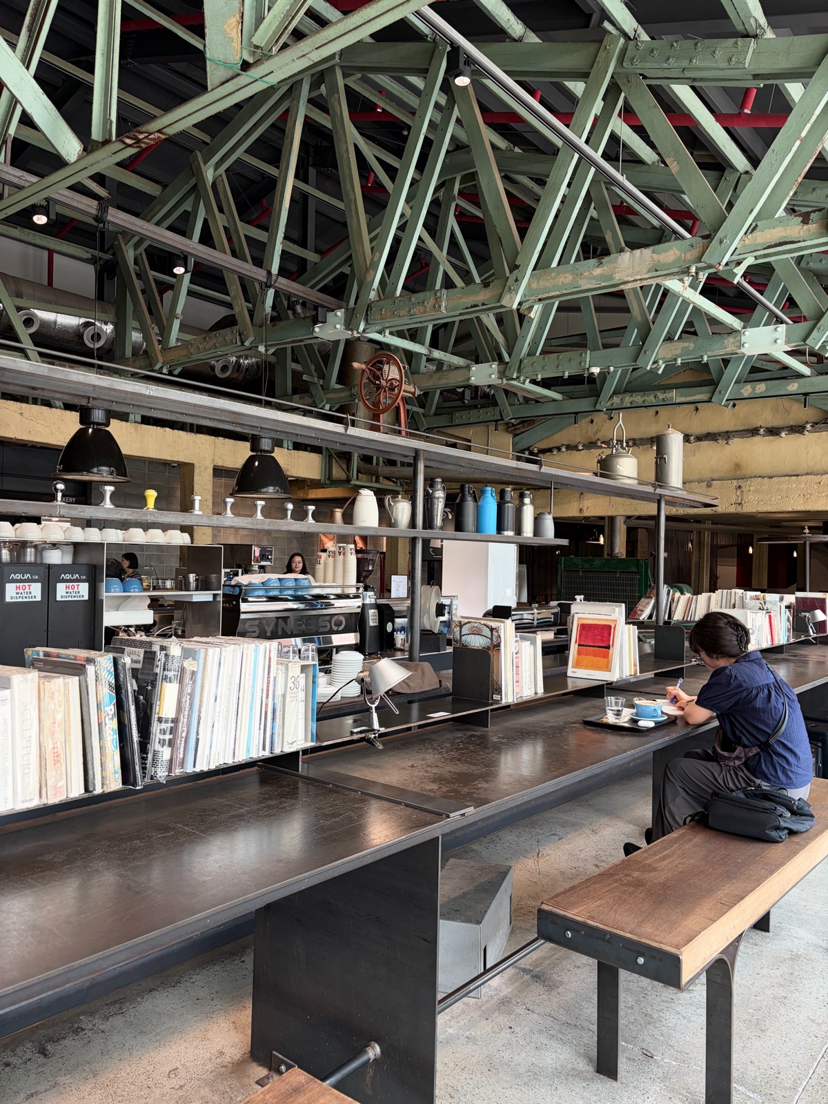 Interior view of a cafe with long tables and books beneath a distinctive green steel-frame ceiling. 독특한 녹색 철골 구조의 천장 아래 긴 테이블과 책들이 놓여 있는 카페 내부 전경.